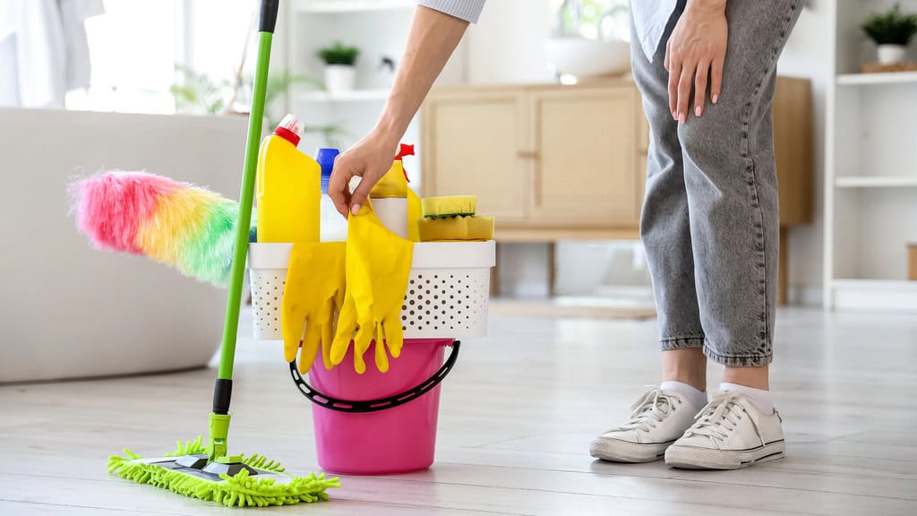 A woman reaching down to a bucket of cleaning products, rubber gloves and a mop, as she's preparing to vacate and clean her home.